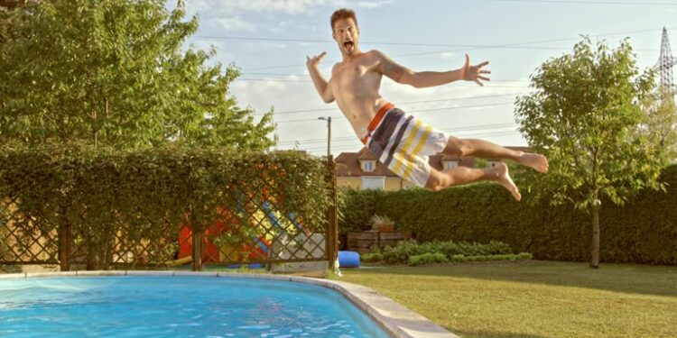 Slow motion medium tracking shot of a young man jumping into the pool in a funny way, making a big splash.